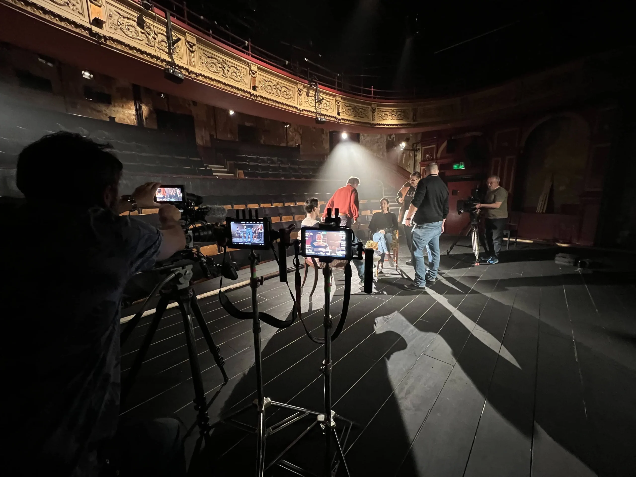 Film crew filming a scene on a dimly lit theater stage, with cast seated beneath a spotlight and ornate balcony above