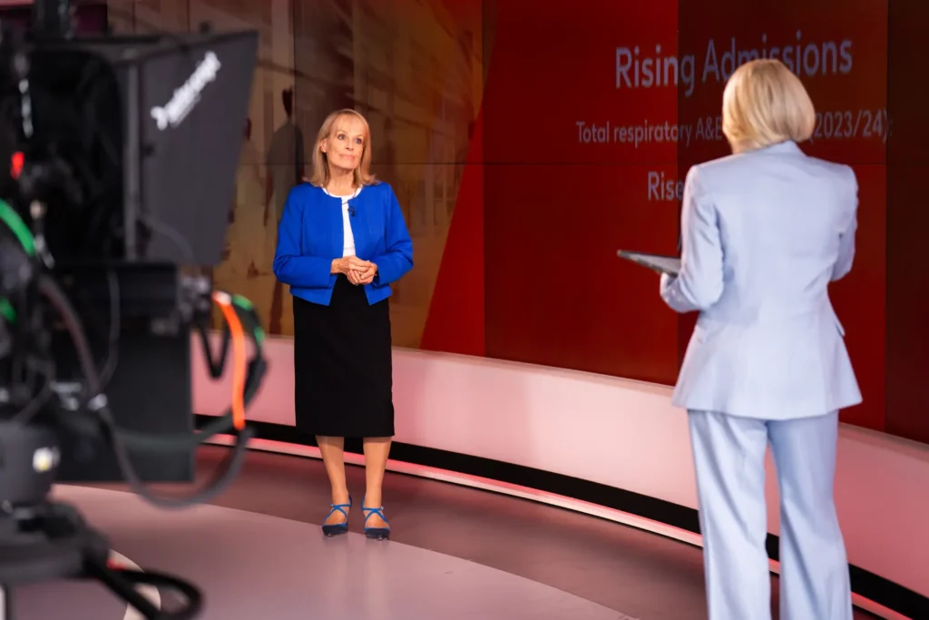Presenter discussing "Rising Admissions" respiratory statistics on a red TV studio backdrop, filmed by broadcast camera
