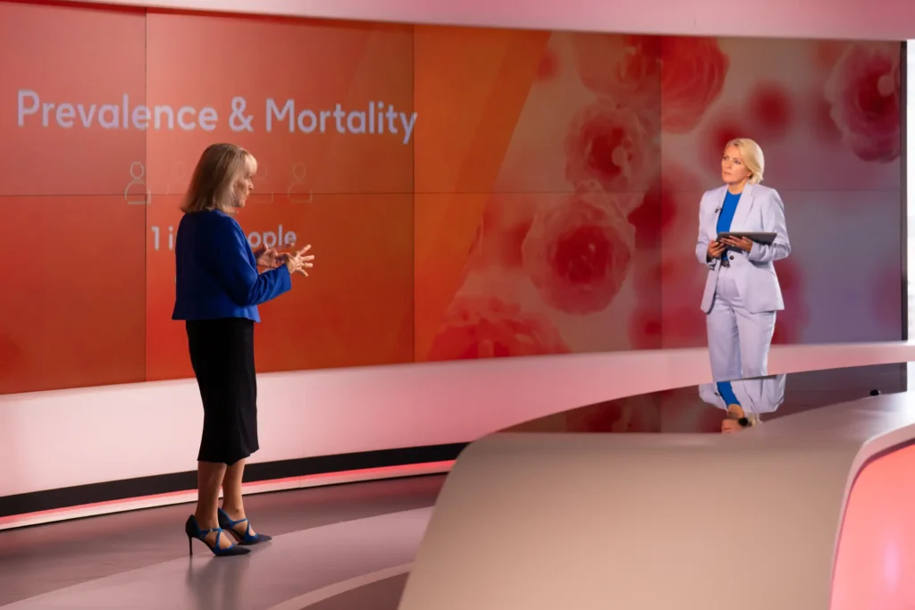 Two presenters on a health news studio set discussing "Prevalence & Mortality" data with cell imagery on screen