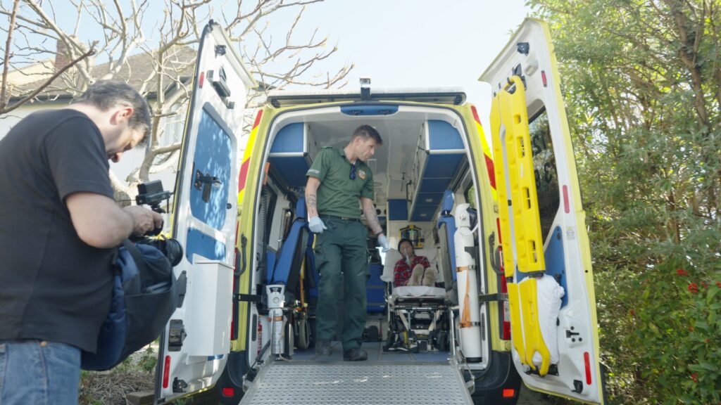 Cameraman filming inside an open ambulance with paramedic assisting a patient on a stretcher