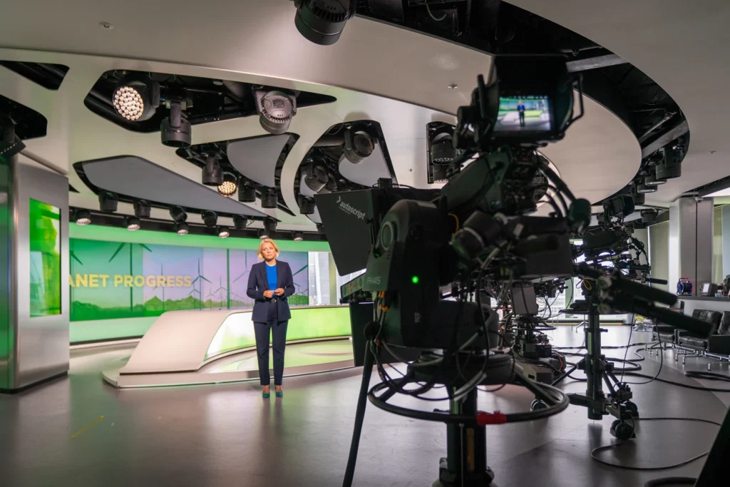 TV news presenter delivering a segment on a green-lit studio set with "Planet Progress" backdrop, camera crew filming